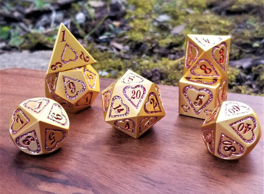 A close-up of a gold metal dice set with pink heart engravings on the faces, placed on a wooden surface with a blurred natural background.