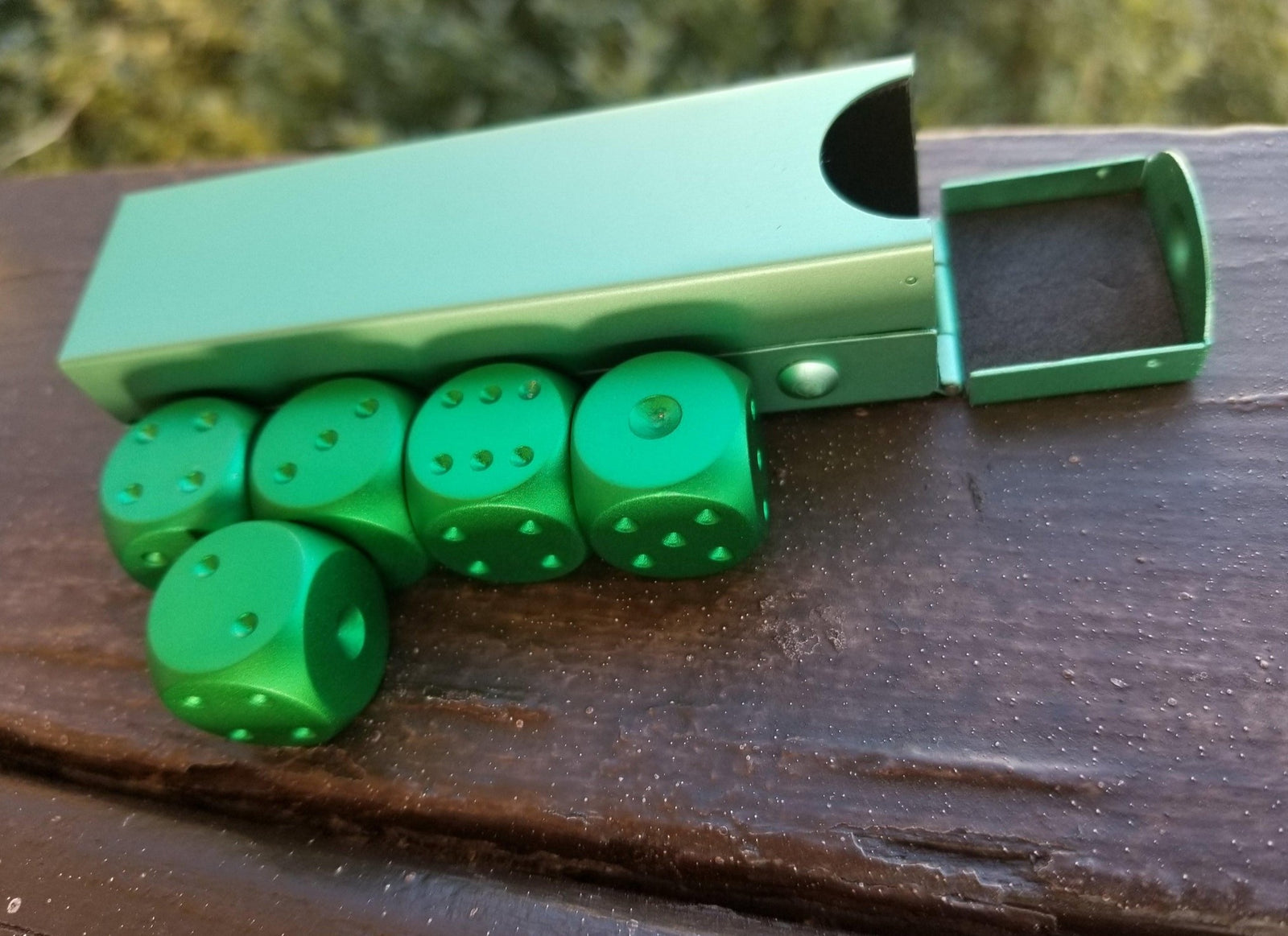 A set of green aluminum D6 dice with pips, displayed alongside a foam-lined case.