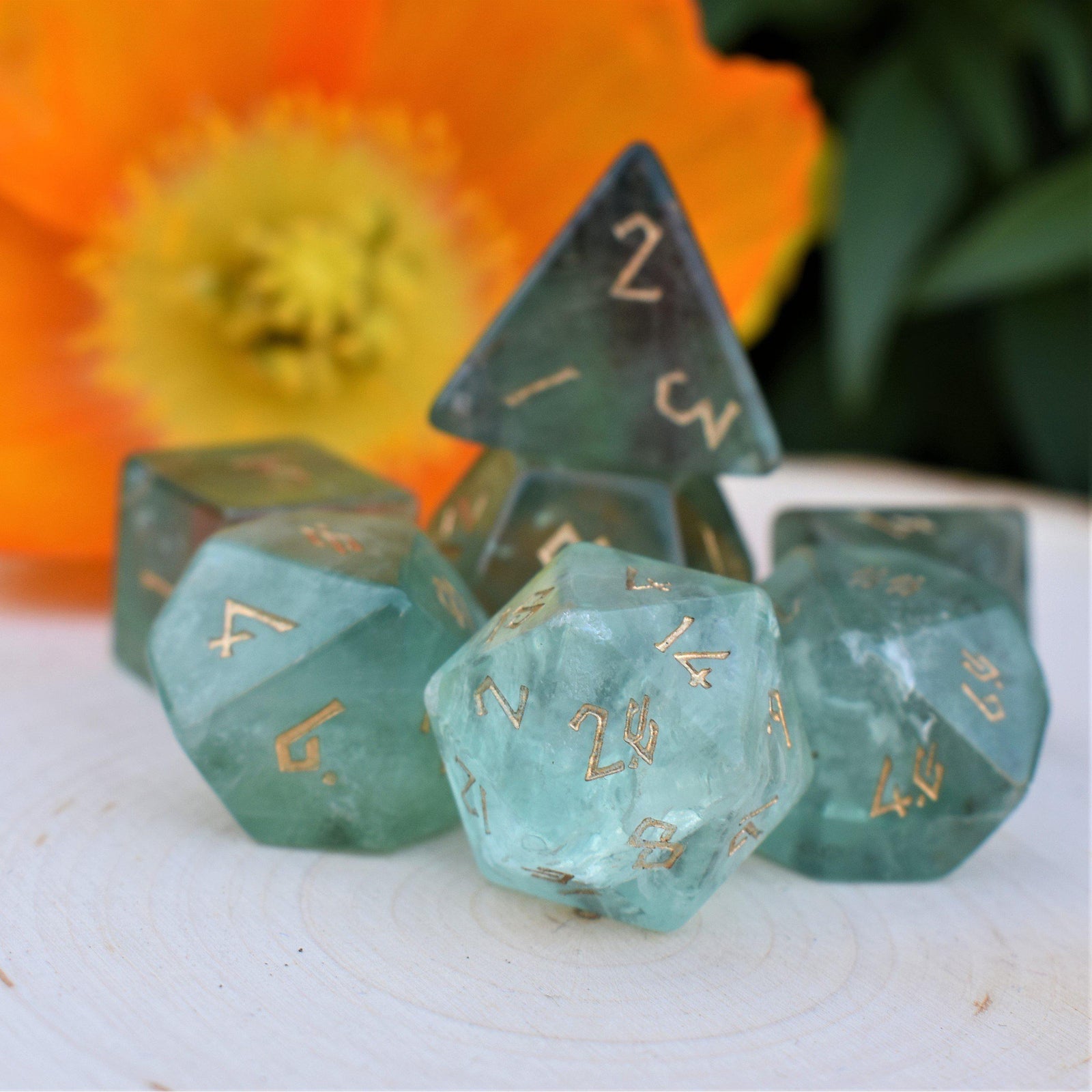 A collection of green fluorite stone dice with engraved numbers, including a D4, D6, D8, two D10, D12, and D20, displayed on a white surface with yellow flowers in the background.