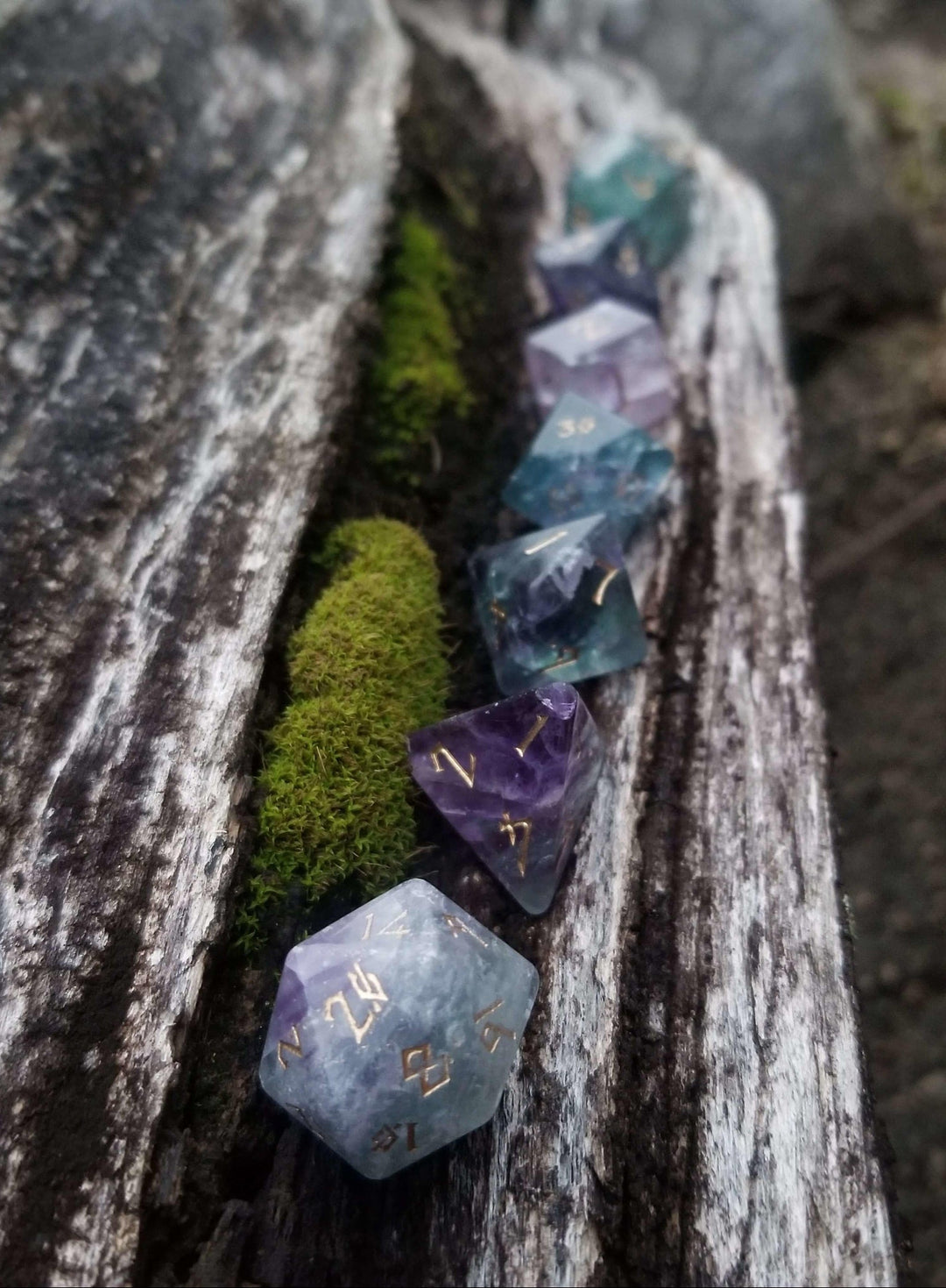 A set of purple fluorite stone dnd dice with engraved numbers, displayed on a wooden surface with flowers in the background.