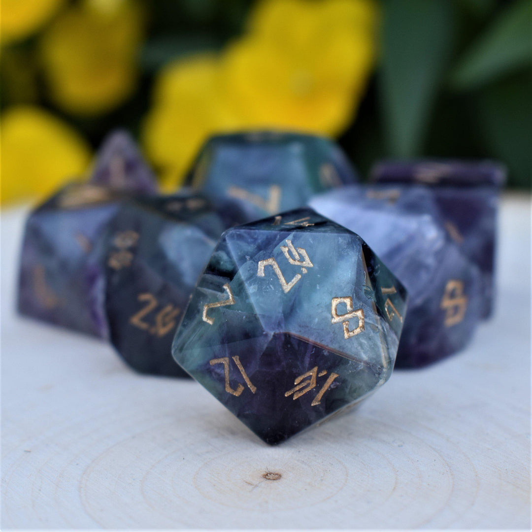 A set of purple fluorite stone dnd dice with engraved numbers, displayed on a wooden surface with flowers in the background.
